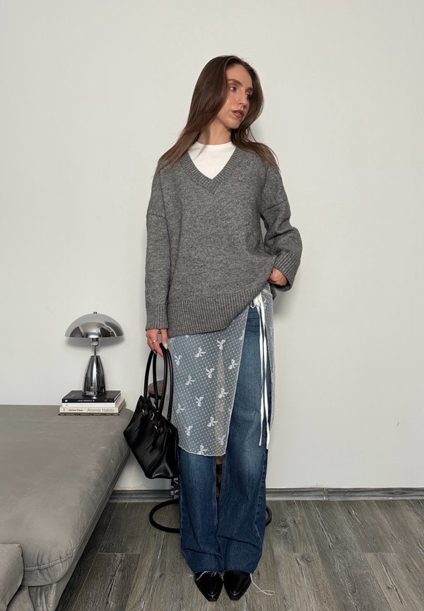 Kvinde iført en grå oversized sweater, hvid top, blå brede jeans, gennemsigtig blomsteroverlay, sorte spidse sko og holder en sort håndtaske.