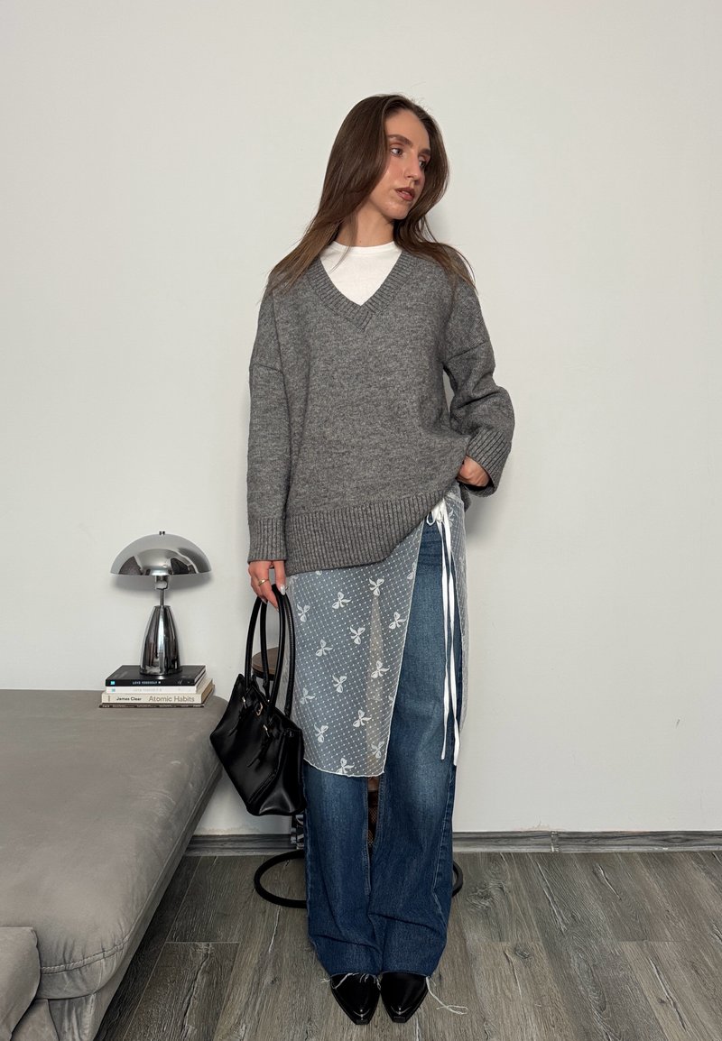 Donna con indosso un maglione grigio oversize, top bianco, jeans a gamba larga blu, sovrapposizione floreale trasparente, scarpe nere con punte appuntite, e che tiene una borsa nera.