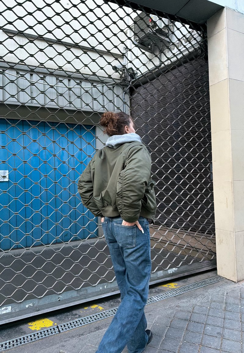 Personne en veste verte et jean, debout les mains dans les poches, face à une porte métallique fermée en milieu urbain.