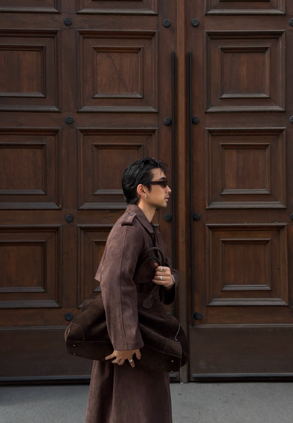 A man wearing sunglasses and a long brown coat is walking past a wooden panelled door, carrying a large matching brown bag.