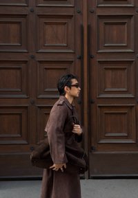 A man wearing sunglasses and a long brown coat is walking past a wooden panelled door, carrying a large matching brown bag.