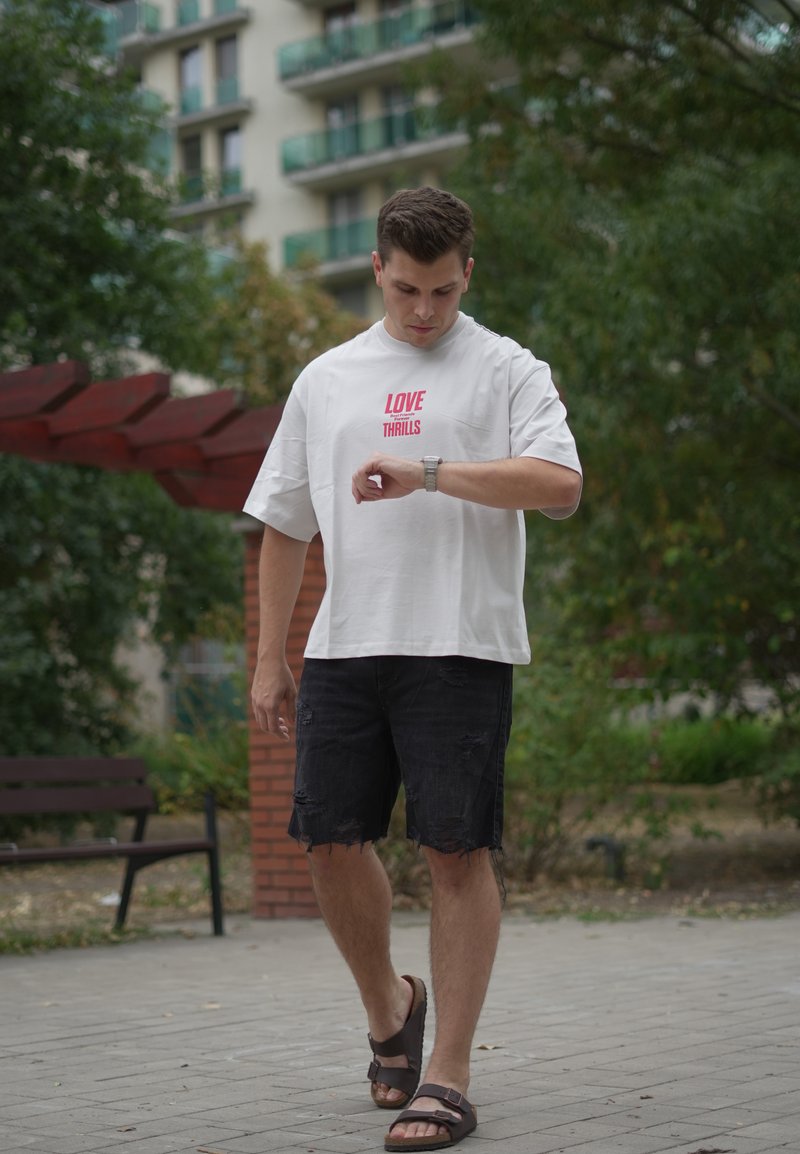 Homme en chemise blanche et short noir marchant dehors, regardant sa montre-bracelet, avec des arbres et un bâtiment en arrière-plan.
