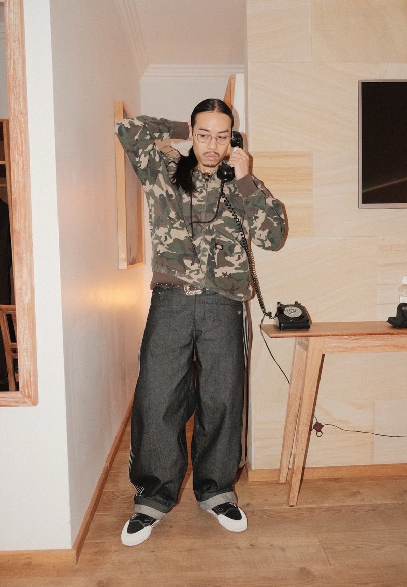 Kapuča z vzorcem za kamuflažo z žepi, temne ohlapne jeans hlače z belimi črtami in črni supergi. Črni vrtljivi telefon na lesenem mizi.