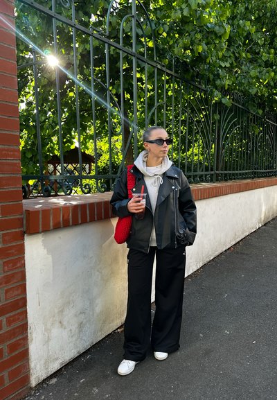 Chaqueta de cuero negra sobre una sudadera gris, combinada con pantalones negros de pierna ancha y zapatillas blancas. Sosteniendo una bebida con una pajilla roja y un bolso.