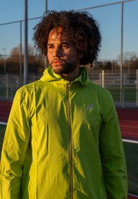 Chaqueta con capucha de color verde brillante con cremallera completa, confeccionada con material ligero. Presenta un logo en el pecho y una textura suave.