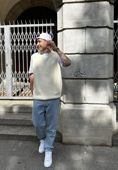 Chaleco de punto beige claro con escote en V sobre una camiseta blanca, combinado con jeans azul claro y zapatillas blancas, de pie junto a un muro de piedra.