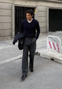 Maglione blu scuro a maniche lunghe sopra una camicia bianca; pantaloni grigi plissettati; scarpe di pelle nere; tiene un cappotto scuro, camminando su una strada.