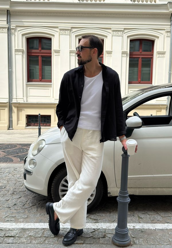 Homme portant des lunettes de soleil, une chemise noire, un pantalon blanc et des chaussures noires, appuyé contre un poteau, tenant une tasse de café à côté d'une voiture blanche.