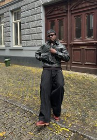 Black leather jacket with a zip and a ribbed collar, teamed with loose black trousers and red chunky shoes. Urban backdrop featuring cobblestones.