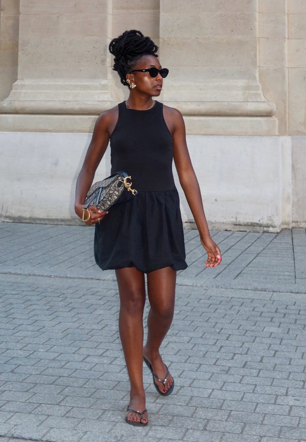 Schwarzes ärmelloses Kleid mit einem schwingenden Rock, kombiniert mit Flip-Flops. Hält eine gemusterte Handtasche. Sonnenbrille und Creolen als Accessoires.