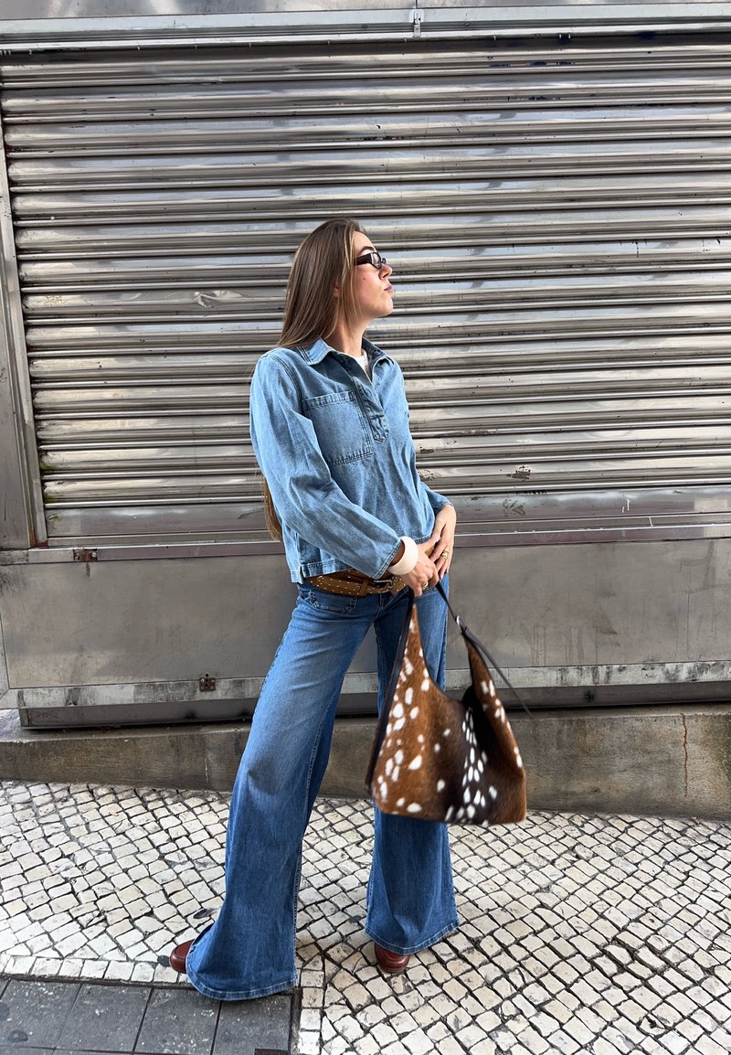 Woman in blue denim outfit and sunglasses holding a brown and white spotted bag standing on a tiled sidewalk by a metal shutter.