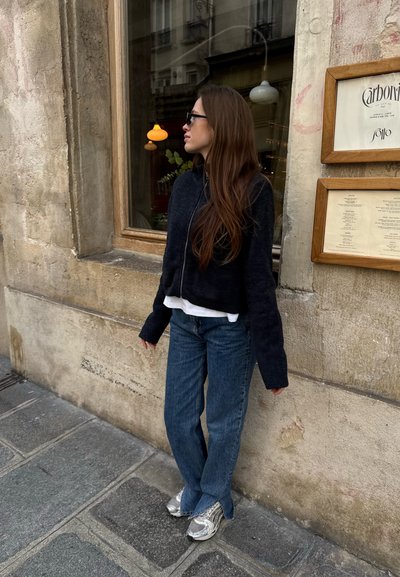 Suéter negro cortado, camisa blanca debajo, jeans azules con un ligero acampanado y zapatillas plateadas, de pie frente a una pared texturada.