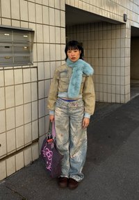 Jeune femme portant une veste en denim usée et un pantalon assorti, une écharpe bleue en fourrure, tenant un sac fourre-tout Hello Kitty violet foncé, debout près d'un mur carrelé en plein air.