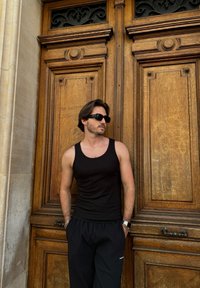 Μαύρο αμάνικο μπλουζάκι και σκούρο παντελόνι μπροστά από μια περίτεχνη ξύλινη πόρτα με σκαλιστές λεπτομέρειες; Γυαλιά ηλίου και διακριτές υφές προσθέτουν αντίθεση.