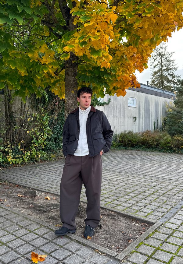 Junger Mann in dunkler Jacke und weiten braunen Hosen steht mit den Händen in den Taschen unter einem gelb-grünen Baum auf gepflastertem Boden neben einem Gebäude.