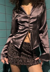 Braune Satinbluse mit Schnürung vorne und langen Ärmeln, kombiniert mit einem Pailletten-Minirock mit strukturiertem Muster.