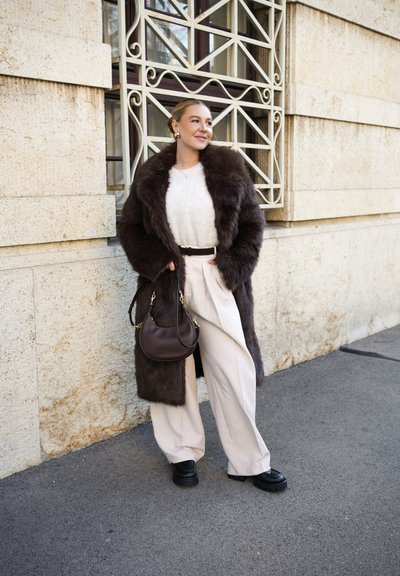 Abrigo de piel marrón, suéter texturizado crema, pantalones beige de pierna ancha, zapatos plataforma negros y un bolso marrón con herrajes dorados. Fondo urbano.
