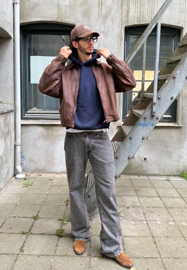 Un hombre con una chaqueta de cuero marrón, una sudadera con capucha azul marino, gorra y vaqueros se ajusta la capucha en el exterior de un edificio, junto a una escalera metálica.