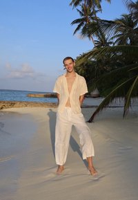 Linen shirt in off-white, buttoned up, with an open collar, paired with loose-fitting white linen trousers, standing on a sandy beach.