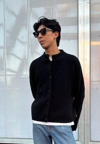 Svart teksturert cardigan med høy krage, frontknapper, båret over en hvit skjorte, sammen med lyseblå jeans og svarte solbriller.