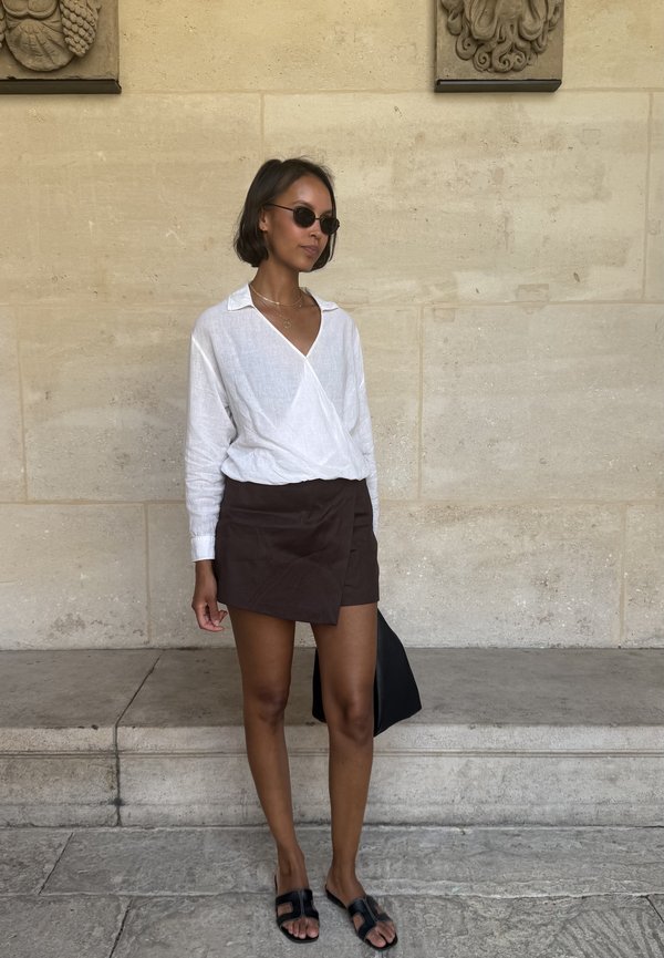 Femme portant des lunettes de soleil rondes, un chemisier blanc croisé, une jupe foncée, des sandales noires, debout contre un mur de pierre tenant un sac noir.