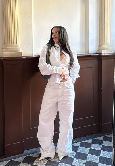 Mujer con cabello largo, vestida con una blusa blanca y pantalones de pierna ancha, de pie en una esquina con paneles de madera oscura y un suelo de baldosas a cuadros.