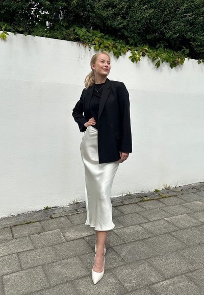 Blazer negro de gran tamaño, falda de satén blanca y zapatos de tacón nude. El fondo presenta una pared blanca y vegetación arriba.