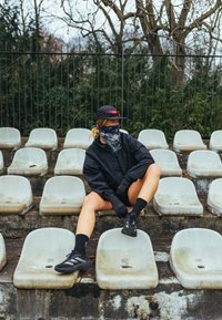 Personne portant une veste noire, des gants, une casquette et un bandana sur le visage, assise sur des sièges d'un stade en plein air usés, avec des arbres et une clôture en arrière-plan.