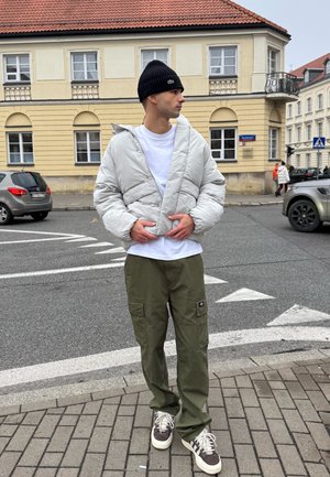 Chaqueta acolchada gris claro, camiseta blanca, pantalones cargo verde oliva y zapatillas marrones. La chaqueta presenta un diseño de superposición único.