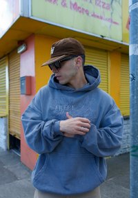 Sudadera gris con diseño impreso "FAVELA Worldwide", corte oversized, puños acanalados. Gorra marrón y gafas de sol. El fondo presenta una pared amarilla brillante y una persiana.