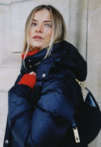 Marinblå vadderad jacka med hög krage, röd stickad tröja under och en liten svart handväska med guldfärgad metall.