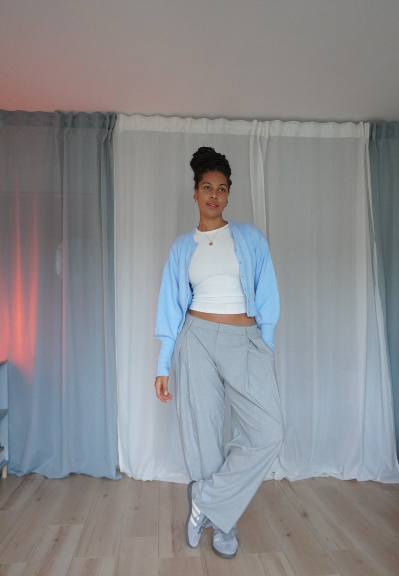 Hellblaues Cardigan über einem weißen Cropped-Top, graue weit geschnittene Hose und graue Sneaker; glatte Texturen, sanfte Farben und lockere Passform.