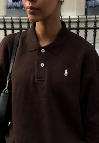 Polo marron avec un col court, trois boutons et un logo blanc brodé. Un sac à main noir est porté sur l'épaule.