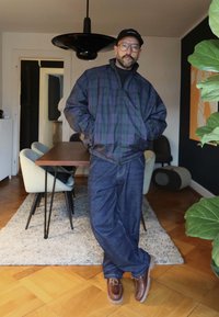 Mann med briller og lue står i spisestuen iført rutete jakke, blå jeans og brune sko, med hendene i lommene, foran et bord og stoler.