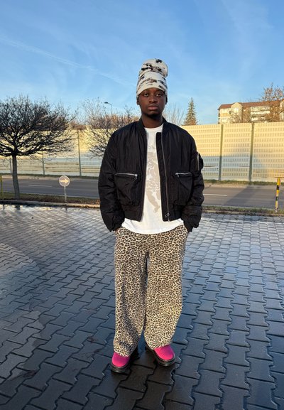 Chaqueta bomber negra, camiseta blanca con diseño gráfico, pantalones de pierna ancha con estampado de leopardo y zuecos rosas. Tejido texturizado y ajuste holgado en todo el conjunto.