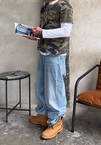 Kamuflāžas T-krekls virs balta garām piedurknēm krekliem, gaiši zila brīvi pieguloša džinsu bikses un brūni zābaki ar auklām un teksturētu zoli. Tur rokā žurnālu.