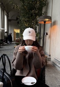 Biely flísový baseballový šilt s čiernym logom, béžový sveter, drží biely pohár, čierna taška, okrúhly tanier so striebornou lyžičkou, vonkajšie prostredie.