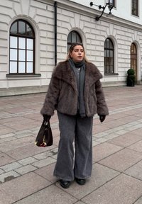 Brun konstläderjacka, grå turtleneck, vida grå jeans, svarta ankelstövlar. Håller en mörk väska med en körsbärscharms. Murad bakgrund.