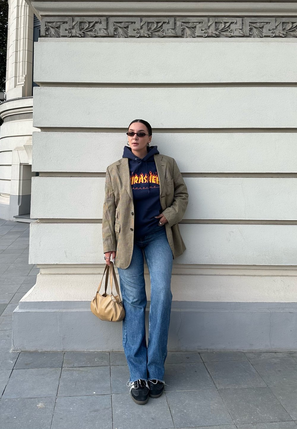 Διπλό αθλητικό σακάκι με δομή σε ανοιχτό καρό, ναυτικής απόχρωσης hoodie με λογότυπο, φαρδές μπλε τζιν, μαύρα sneakers και μπεζ τσάντα, ποζάροντας μπροστά σε τοίχο.