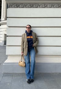 Διπλό αθλητικό σακάκι με δομή σε ανοιχτό καρό, ναυτικής απόχρωσης hoodie με λογότυπο, φαρδές μπλε τζιν, μαύρα sneakers και μπεζ τσάντα, ποζάροντας μπροστά σε τοίχο.