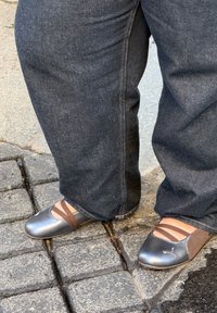 Chaussures gris satin avec des lanières brunes sur le cou-de-pied, bout arrondi et marquage discret, portées avec un jean bleu foncé sur un sol pavé.
