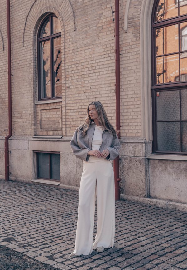 Vrouw met lang haar die een grijze korte jas en brede witte broek draagt, staat op een kasseienstraat naast een bakstenen gebouw met ramen.