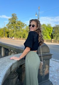 Black cropped top and light green wide-leg pants, standing by a stone railing, with sunglasses and a small chain handbag. Outdoor setting.