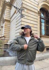 Olive green bomber jacket with a fur-lined hood, zip front, and ribbed cuffs. Worn over a white shirt, paired with grey trousers.