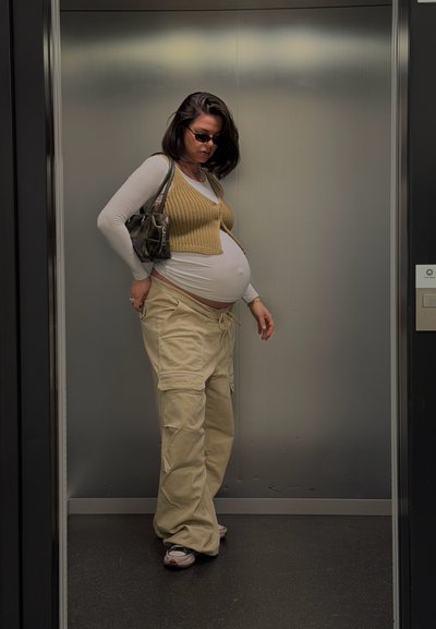 Mujer embarazada con gafas de sol, pantalones cargo beige, camiseta blanca de manga larga y chaleco de punto, de pie dentro de un ascensor con un bolso.