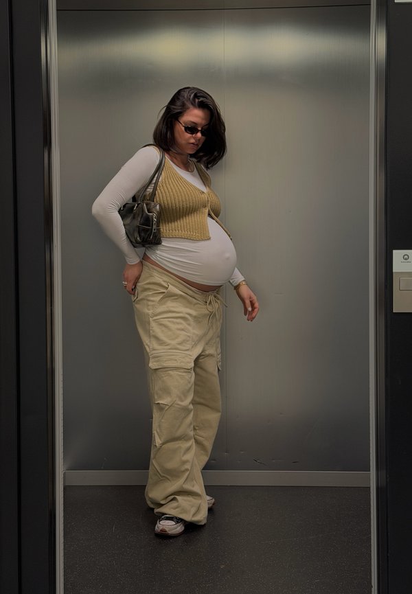 Mujer embarazada con gafas de sol, pantalones cargo beige, camiseta blanca de manga larga y chaleco de punto, de pie dentro de un ascensor con un bolso.