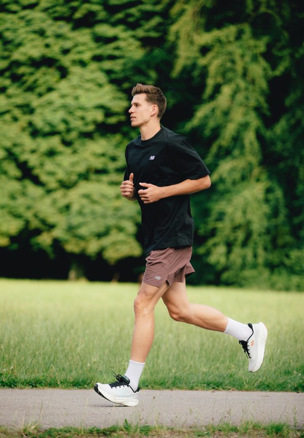 Corredor masculino con una camiseta negra de manga corta y pantalones cortos marrones, vistiendo zapatillas de correr negras y blancas, trotando por un sendero rodeado de hierba.