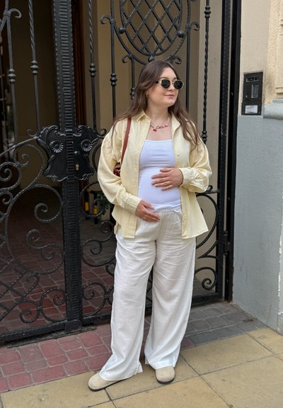 Camisa amarilla clara de botones sobre una camiseta blanca, combinada con pantalones blancos holgados y zapatos beige; de pie frente a una puerta negra ornamental.