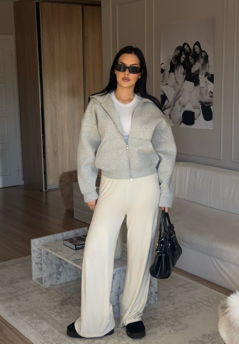 Sudadera gris con cremallera, camiseta blanca, pantalones anchos crema, gafas de sol negras y bolso. Texturas suaves; paleta de colores minimalista.