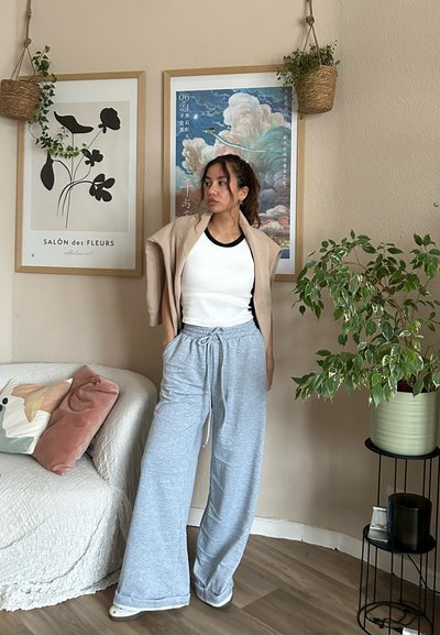 Pantalones de chándal gris de pierna ancha, top blanco sin mangas y un cárdigan oversize beige. La habitación cuenta con obras de arte en las paredes, un sofá texturizado y una planta verde.